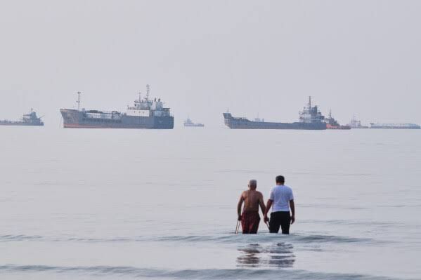Two men stand in the waters of the Strait of Hormuz with anchored vessels visible in the distance off Bandar Abbas, Iran.