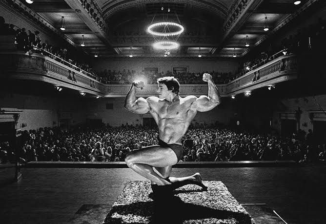 A vintage 1975 photograph of Arnold Schwarzenegger peeking through a stage curtain, now held by the MoMA.