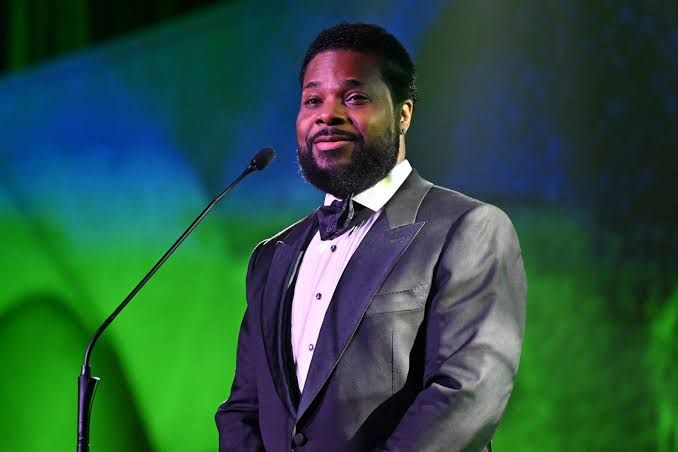 Malcolm-Jamal Warner stands in a suit, representing the subject of an estate lawsuit between his widow and mother.