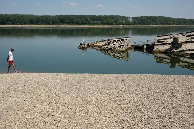 A man stands on a sandy riverbank near the exposed hull of a German warship from World War II on the Danube River.