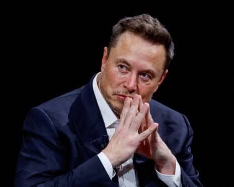 Elon Musk presses his fingertips together thoughtfully near his chin while speaking at a public event.