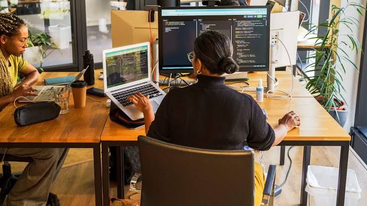 A developer working on code at a dual-monitor workstation next to a colleague.