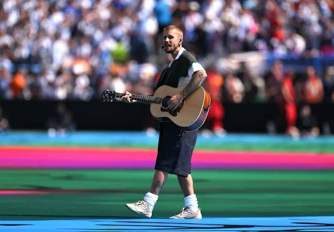 Justin Bieber performing with an acoustic guitar at the center of the FIFA World Cup Final stadium pitch.