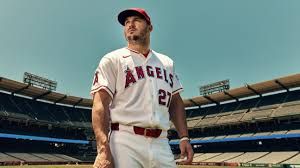 Mike Trout standing in an Angels dugout, looking thoughtful while holding a baseball bat.