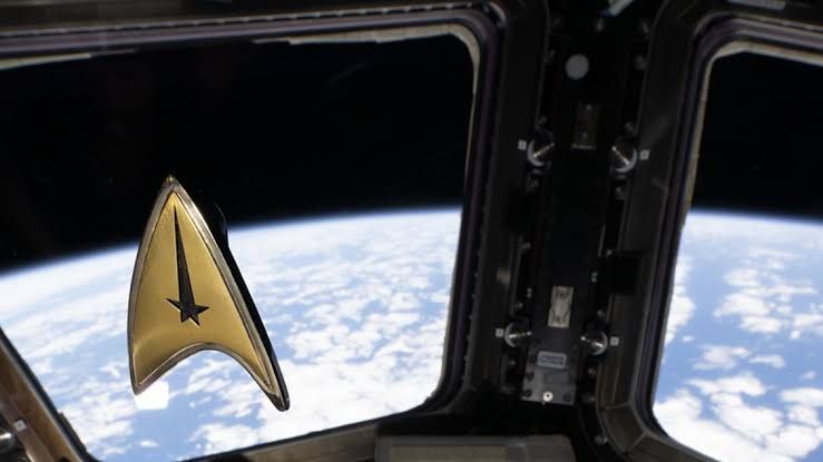 Astronaut Frank Rubio floats in the ISS Cupola module holding a Star Trek anniversary emblem.