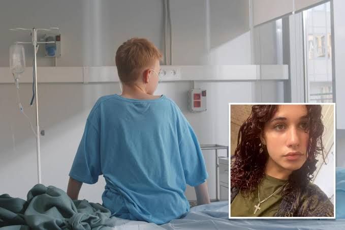 A preteen boy in a hospital gown sits on a bed and looks out a window.
