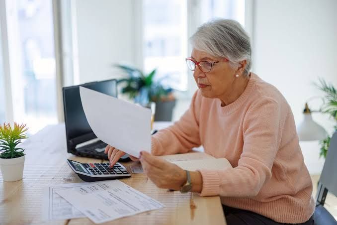 A retiree sits at a desk while using a calculator to review financial documents.