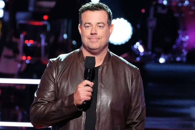 Carson Daly speaking into a handheld microphone on the set of The Voice.