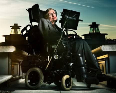 Stephen Hawking in his wheelchair at a public lecture, appearing before a large screen displaying complex physics equations.