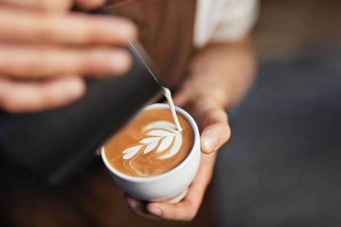 A smiling young man wearing a barista apron prepares coffee at a cafe counter in a professional hospitality setting.