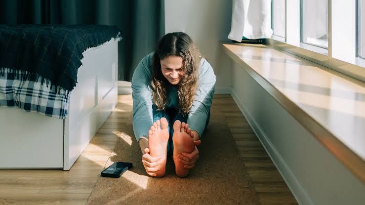 A woman performing a forward fold yoga pose on a mat in a bedroom to stretch her hamstrings.
