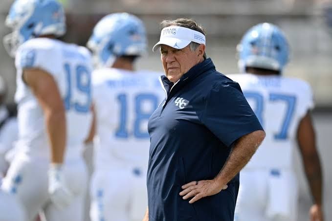 Bill Belichick stands on the sidelines at Kenan Stadium during a UNC football game with empty seats visible.