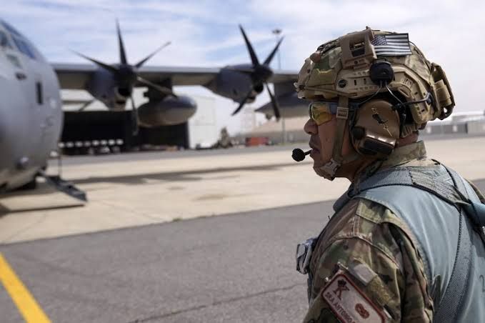 A U.S. service member wearing a helmet stands on an airfield in Jordan near a large military transport plane.