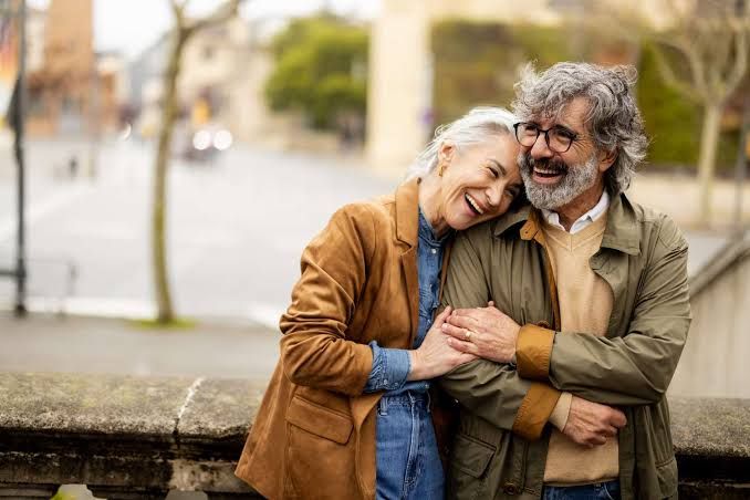 An elderly couple smiling and embracing outdoors in a park setting.