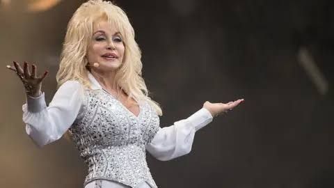 Dolly Parton wearing a white and silver bedazzled outfit smiling on stage while holding out her arms.