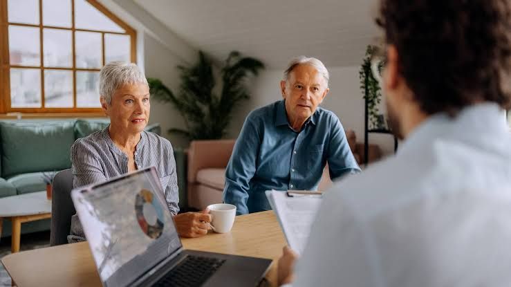 An elderly couple reviews retirement financial documents and Medicare statements at a kitchen table.