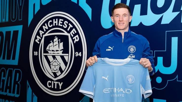 Manchester City midfielder Elliot Anderson poses with his new team shirt in front of a club-branded wall.