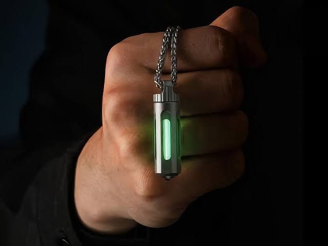 A close-up of a titanium keychain cylinder glowing green on a desk next to other small metal tools.