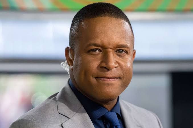 Craig Melvin sitting at the Today show desk in Studio 1A wearing a suit.