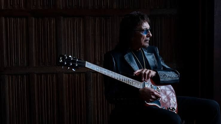 Tony Iommi playing a Gibson SG guitar during a live performance in a dark stage setting.