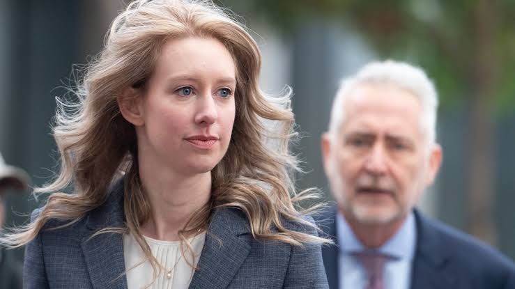 Elizabeth Holmes standing in court during a hearing, wearing a dark blazer, reflecting her legal situation.