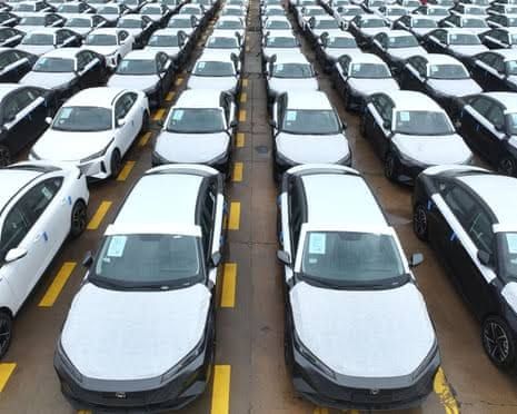 Rows of white cars with yellow wheel blocks parked in a large distribution lot in China.