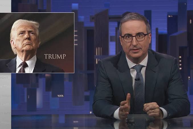 John Oliver sitting at his desk on the set of Last Week Tonight with a graphic of Donald Trump behind him.