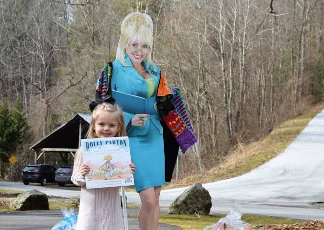 A young child holds a book by Dolly Parton in front of a cardboard cutout of the singer.