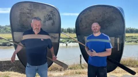 Andrew Davies and Keith Elias standing with their traditional coracles on the banks of the polluted River Tywi.