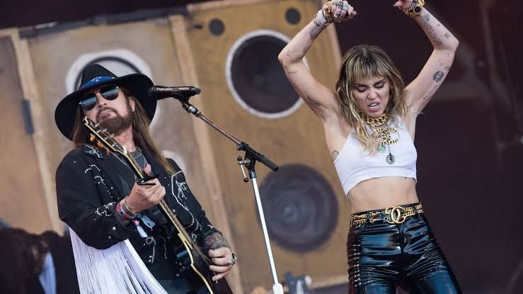 Billy Ray Cyrus playing guitar while Miley performs on stage during a concert.