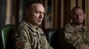 Major General Mykhailo Drapatyi sits in his military uniform while an associate observes in the background.