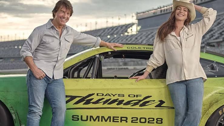 Tom Cruise and Anne Hathaway standing in front of a modern NASCAR vehicle at a track.