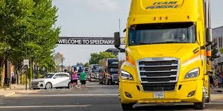 A fleet of colorful NASCAR haulers driving down a street lined with fans in the town of Speedway, Indiana.