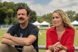 Jason Sudeikis and Andrea Anders sit on the grass by a football pitch during the filming of Ted Lasso season 4.