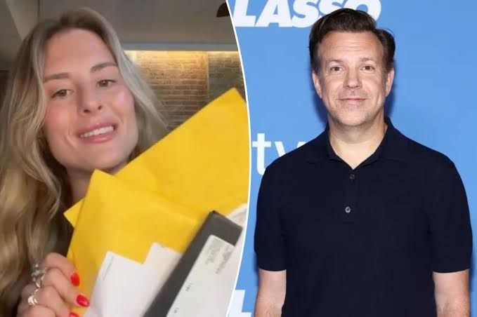 Jason Sudeikis smiling on a red carpet while holding a stack of mail handed to him by Taylor Palmby at a movie premiere.