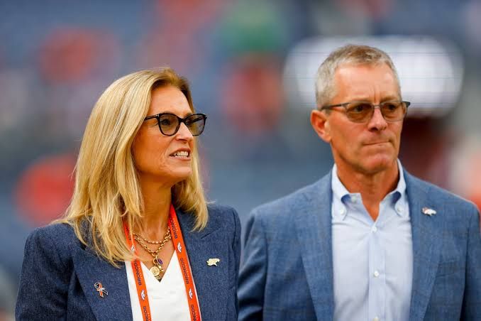 Carrie Walton Penner and Greg Penner walking on the sidelines of an NFL stadium during a Broncos game.
