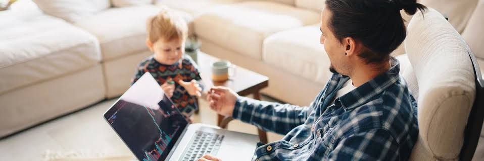 A man working on a laptop with financial charts displayed while his young child watches in a home office setting.