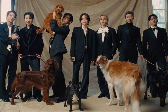 BTS members standing in a row facing away from the camera in front of a bank of bathroom urinals.