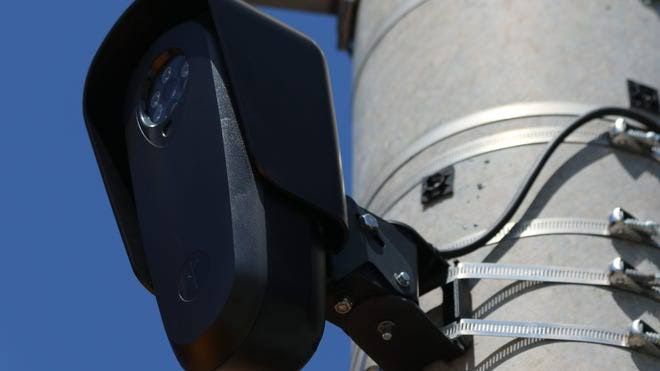 Flock license plate reader camera mounted on a metal pole above a city intersection.
